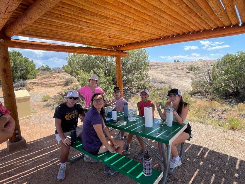 A group of people sitting under an awning at a picnic table having lunch.